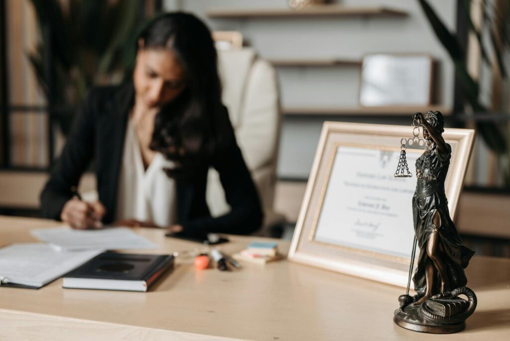 Focused woman writing at desk with Lady Justice figurine and certificate in office.